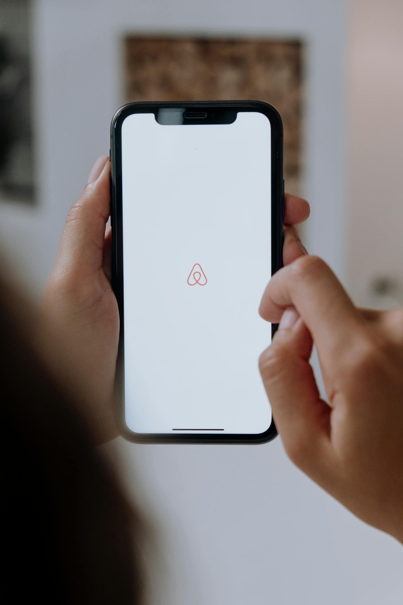 A person holding an iPhone with the Airbnb app logo on screen, representing Airbnb's mobile-first travel marketplace.