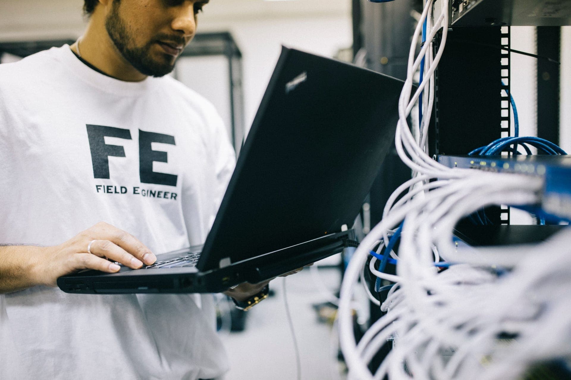 A field engineer using a laptop while inspecting network cables and hardware in a server room