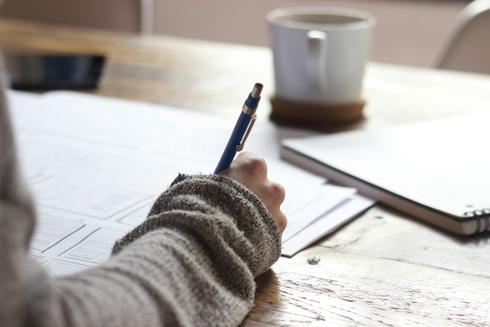 A person writing notes at a wooden table beside a white coffee mug, representing the careful customization step that turns a generic cover letter template into a job-specific draft.