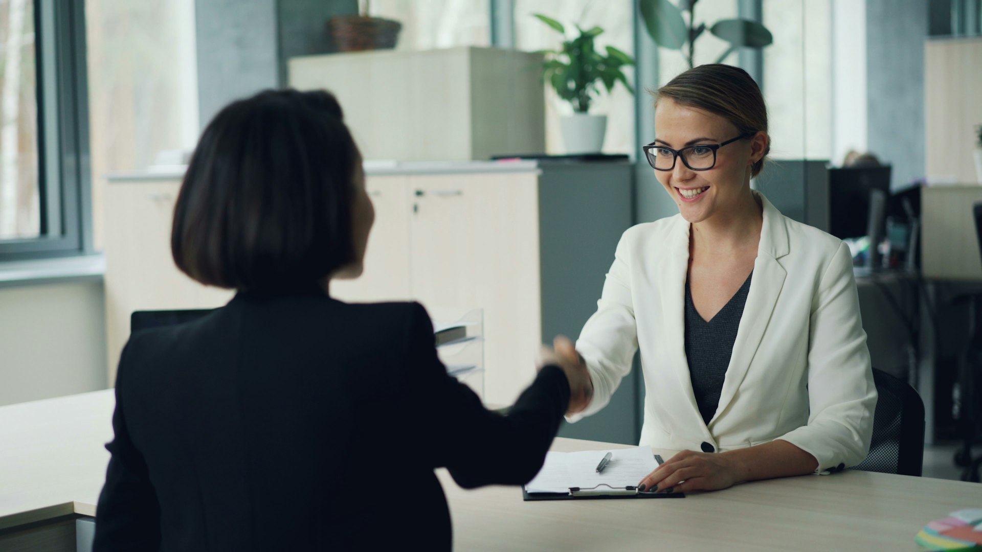 Two professionals in smart-casual attire shaking hands across a desk during an interview.