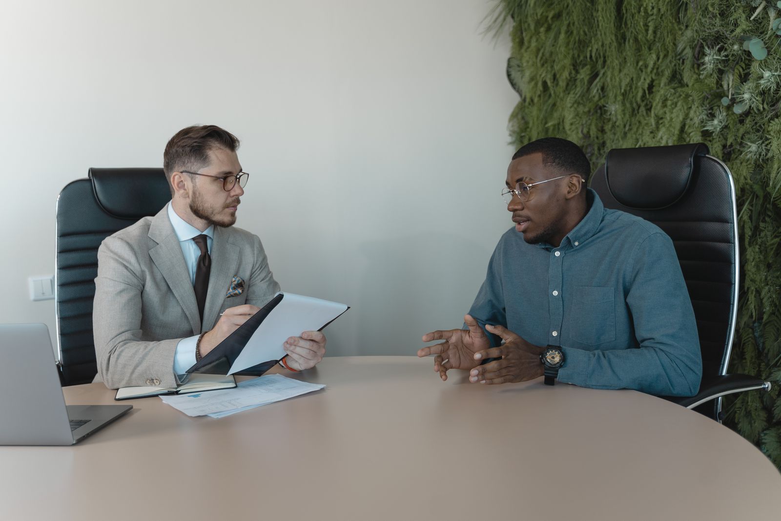 Two professionals talking across a conference table during a job interview, one holding a resume.